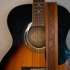 Puede incluir: Una correa de guitarra de cuero marr&oacute;n con un dise&ntilde;o floral. La correa est&aacute; unida a una guitarra ac&uacute;stica negra y marr&oacute;n con el nombre de la marca Mitchell.