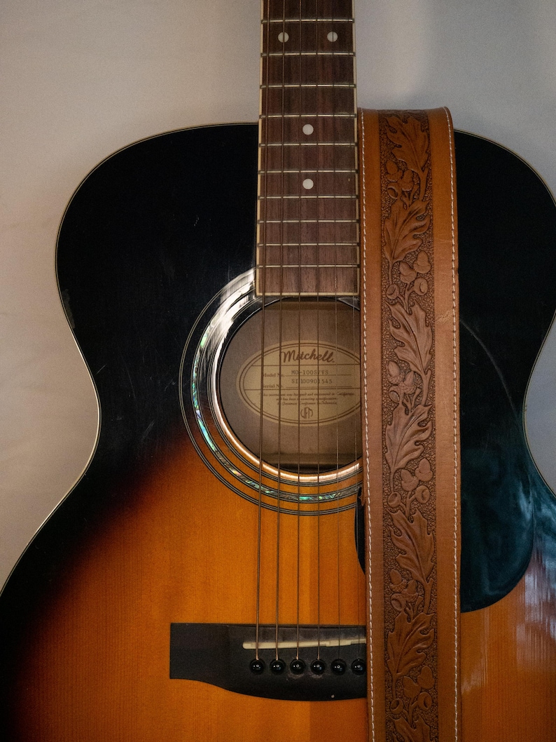 Puede incluir: Una correa de guitarra de cuero marr&oacute;n con un dise&ntilde;o floral. La correa est&aacute; unida a una guitarra ac&uacute;stica negra y marr&oacute;n con el nombre de la marca Mitchell.