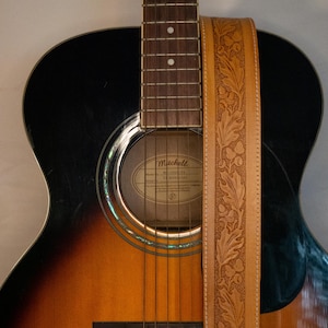 Puede incluir: Una guitarra ac&uacute;stica negra y marr&oacute;n con una correa de guitarra de cuero con un dise&ntilde;o de hojas en relieve. La guitarra tiene una boca de sonido con una etiqueta que dice "Mitchell" y "Modelo 3200113".