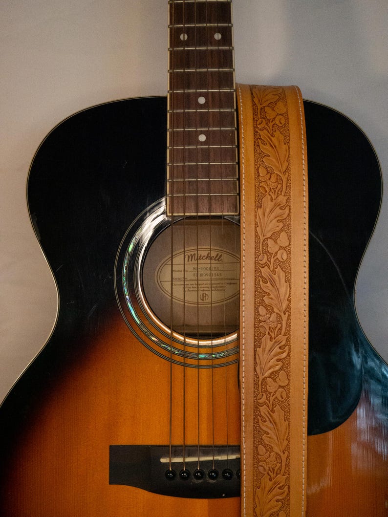Puede incluir: Una guitarra ac&uacute;stica negra y marr&oacute;n con una correa de guitarra de cuero con un dise&ntilde;o de hojas en relieve. La guitarra tiene una boca de sonido con una etiqueta que dice "Mitchell" y "Modelo 3200113".