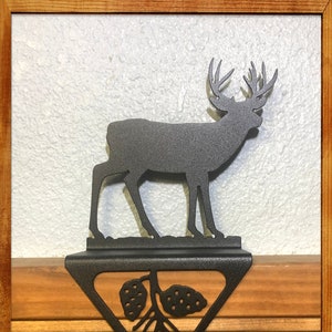 May include: Black metal stocking holder featuring a silhouette of a deer with antlers and pine cones. The holder is attached to a wooden mantel.