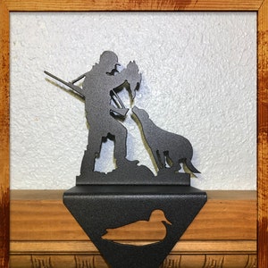 May include: Black metal stocking holder featuring a silhouette of a hunter with two dogs and a duck. The hunter is holding a gun and a duck. The stocking holder is shaped like a triangle with a duck silhouette at the bottom.