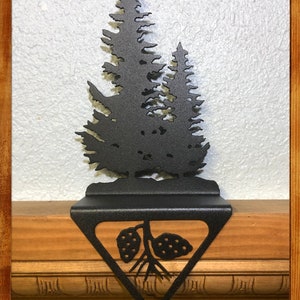 May include: Black metal stocking holder shaped like a pine tree with a pine cone on the bottom. The stocking holder is mounted on a wooden surface.
