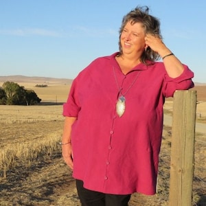 May include: A woman wearing a long-sleeved, magenta button-down shirt with a silver feather pendant necklace. She is standing in a field with a wooden fence post behind her.