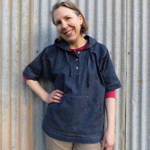 Op de afbeelding: Een vrouw draagt een donkerblauw denim shirt met capuchon, met een enkele knoopsluiting en een klepzak. Het shirt heeft korte mouwen en een losse pasvorm. Ze staat voor een gegolfde metalen muur.