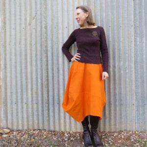 May include: A woman wearing a brown sweater with a broach and an orange skirt. She is standing in front of a corrugated metal wall.
