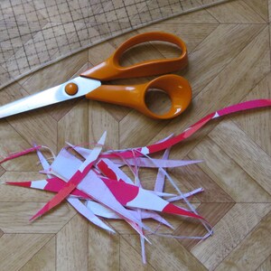 May include: Orange handled scissors with a pile of pink and white fabric scraps on a wood grain surface.