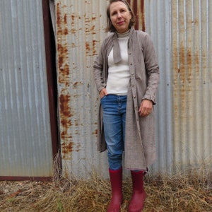 May include: A woman wearing a long brown and white plaid shirt dress with a white turtleneck underneath. She is wearing blue jeans and red rain boots. She is standing in front of a corrugated metal wall.