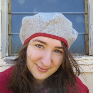 May include: A gray herringbone beret with a red trim. The hat is made of a soft fabric and has a classic, round shape. The beret is a stylish accessory for any outfit.