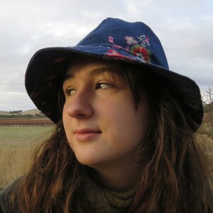 May include: A woman wearing a blue denim hat with floral embroidery. The hat has a wide brim and is tilted slightly to the side. The woman has long brown hair and is looking off to the side.