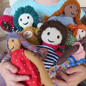 May include: A collection of handmade fabric dolls with different hair colors and clothing. The dolls are all smiling and have embroidered faces. The dolls are being held by a person's hands.