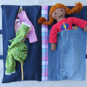 May include: A blue denim travel bag with a zipper closure. The bag has a pink and blue plaid fabric lining and a pocket for a doll. A doll with red hair and a red shirt with a lightning bolt design is in the pocket.