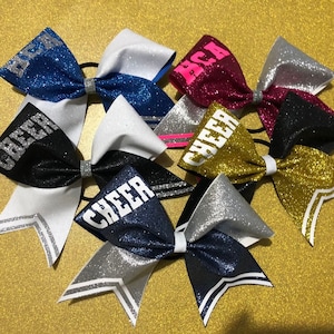 Può includere: Cinque fiocchi da cheerleader con glitter e strisce bianche. I fiocchi sono di colore nero, blu, oro, argento e bordeaux. I fiocchi hanno le parole "Cheer" e "BCA" scritte su di essi.