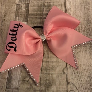 May include: A pink cheer bow with a black embroidered name "Dolly" and silver rhinestones on the edges.