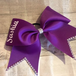 May include: Purple cheer bow with rhinestones and the name "Saskia" written in white.
