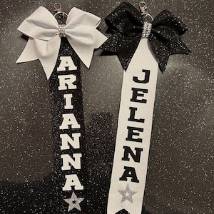 May include: Two personalised cheerleading ribbons with glittery bows. One ribbon is black with a silver bow and the name "ARIANA" in white block letters. The other is white with a black bow and the name "JELENA". Each has a star.