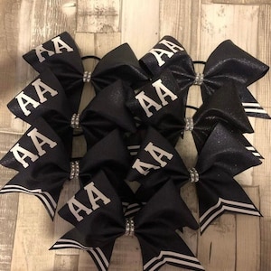 Peut inclure: Six nœuds de cheerleaders noirs avec des rayures blanches et des strass argentés. Chaque nœud porte les lettres "AA" en blanc au centre.