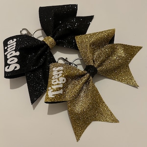 May include: Three glittery cheerleading bows in black and gold. One black bow has the name "Sophie" in white. Another gold bow has the word "Tigers" in white. Each bow has a keychain clip.