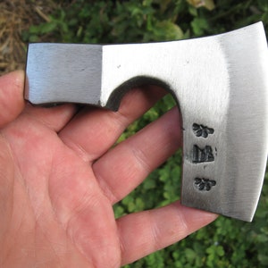 Puede incluir: Un pequeño hacha de color plateado con una hoja curva y un mango de madera. El hacha tiene un pequeño diseño grabado en la hoja.