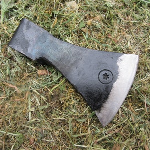 Puede incluir: Una cabeza de hacha negra con un borde ligeramente redondeado y una pequeña marca en forma de estrella en el lateral. La cabeza del hacha está sobre un lecho de hierba seca.