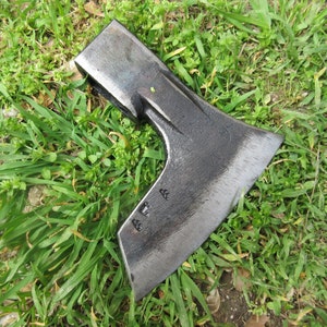 Könnte beinhalten: Eine schwarze Axt aus Metall mit einer leicht gebogenen Klinge. Die Axt hat einen Holzstiel und liegt auf einem Bett aus grünem Gras.