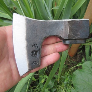 Puede incluir: Una mano sostiene un pequeño hacha forjada a mano con una hoja pintada de blanco y un mango negro. El hacha tiene una pequeña marca decorativa en la hoja.