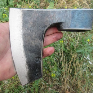 Op de afbeelding: Een hand houdt een zwarte en zilveren bijl vast met een gebogen blad. De bijl heeft een klein, rechthoekig gat in de kop.