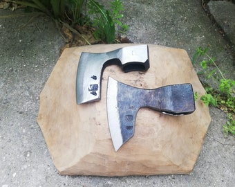 Peut inclure: Deux haches forgées à la main avec des manches en bois, reposant sur une surface en bois. Les haches ont une finition argentée et sont marquées de la lettre "V".