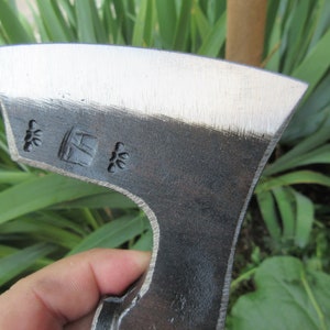 Puede incluir: Primer plano de una cabeza de hacha negra con una hoja plateada. La cabeza del hacha tiene una peque&ntilde;a marca cuadrada con las letras "H" y "K" dentro, y dos peque&ntilde;as marcas en forma de estrella a cada lado de la marca cuadrada.