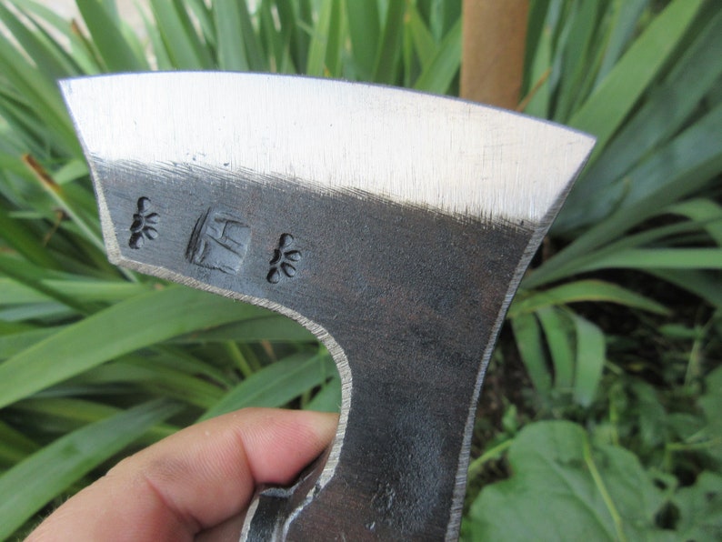 Puede incluir: Primer plano de una cabeza de hacha negra con una hoja plateada. La cabeza del hacha tiene una peque&ntilde;a marca cuadrada con las letras "H" y "K" dentro, y dos peque&ntilde;as marcas en forma de estrella a cada lado de la marca cuadrada.