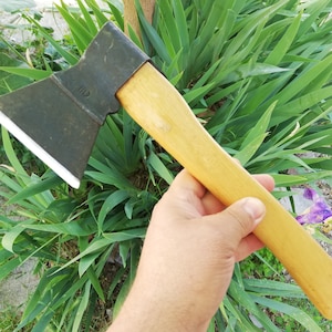 Puede incluir: Un hacha con mango de madera y cabeza de metal negro. El hacha tiene un pequeño agujero en el extremo del mango.