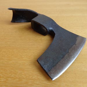 May include: A black metal hand axe with a curved blade and a wooden handle. The axe is on a wooden surface.