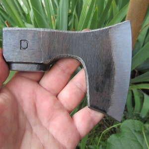 Puede incluir: Un hacha peque&ntilde;a, forjada a mano con un acabado gris oscuro. La cabeza del hacha tiene una ligera curva y un borde redondeado. La cabeza del hacha est&aacute; marcada con la letra "D".