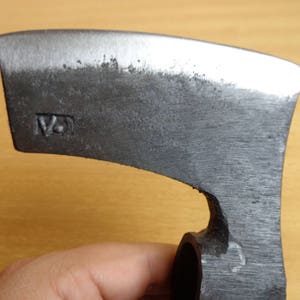 May include: A close-up of a gray metal axe head with a curved blade. The axe head has a small hole in the side and the text "12" is stamped on the side of the axe head.