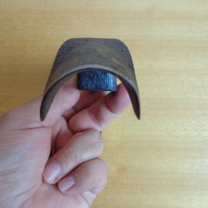 May include: A close-up of a dark brown leather strop with a black wooden handle. The strop is curved and is being held by a hand.