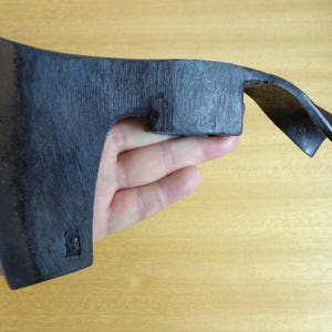 May include: A black hand axe with a curved blade and a wooden handle. The axe is held in a hand, with the blade facing the camera.