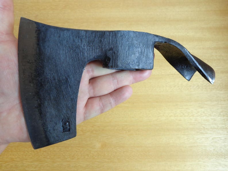 May include: A black hand axe with a curved blade and a wooden handle. The axe is held in a hand, with the blade facing the camera.