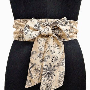 May include: A beige fabric obi belt with a black illustrated pattern of figures, buildings, and palm trees. The belt is tied in a bow at the front, creating a stylish and decorative accessory. The belt is wrapped around a black mannequin.