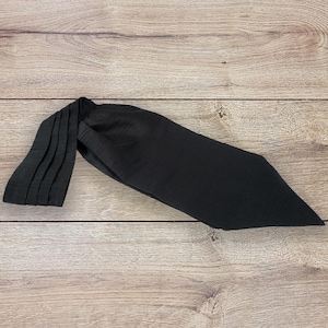 Puede incluir: Una diadema negra de tela suave, con un detalle plisado en un extremo. La diadema está colocada sobre una superficie de madera. La tela tiene un acabado mate.
