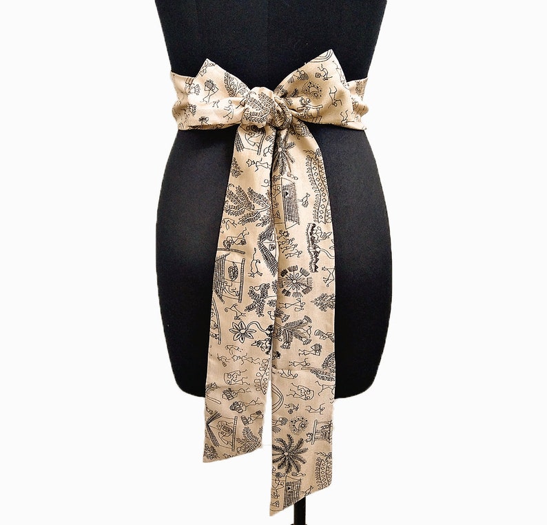 May include: A beige fabric belt tied in a bow, draped on a black mannequin. The belt features a repeating black pattern of stylized figures, plants, and architectural elements. The belt is tied around the mannequin's waist.