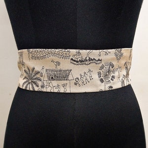 May include: A beige fabric belt with a black tribal pattern, wrapped around a black mannequin. The belt features a repeating design of figures, trees, and geometric shapes. The belt is a wide style, suitable for waist or hip wear.