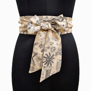 May include: A beige fabric obi belt with a black illustrated pattern of figures, buildings, and palm trees. The belt is tied in a bow at the front, creating a layered effect. The belt is wrapped around a black mannequin.