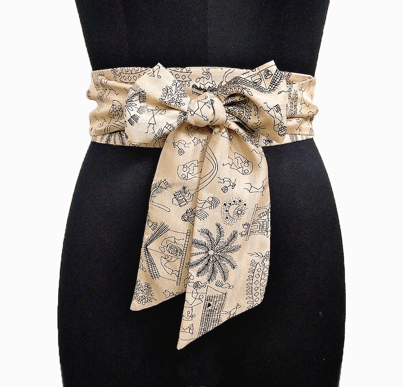 May include: A beige fabric obi belt with a black illustrated pattern of figures, buildings, and palm trees. The belt is tied in a bow at the front, creating a layered effect. The belt is wrapped around a black mannequin.
