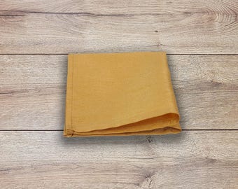 May include: Folded mustard-colored linen napkin. The square napkin is made of a soft, textured fabric. The napkin is folded neatly and is set against a light wood grain background.