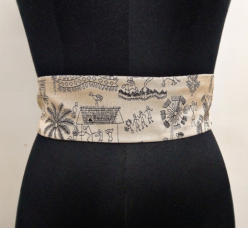 May include: A beige fabric belt with a black tribal pattern. The belt is wrapped around a black mannequin. The pattern includes figures, houses, trees, and geometric shapes. The belt is a wide style.