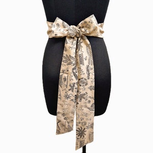 May include: A beige fabric belt tied in a bow, draped on a black mannequin. The belt features a repeating black pattern of stylized figures, plants, and architectural elements. The belt is tied around the mannequin's waist.