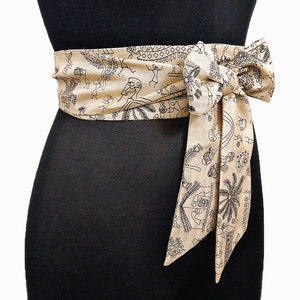 May include: Tan fabric belt with a black tribal pattern tied in a bow. The belt is wrapped around a black mannequin. The fabric features a repeating design of figures, trees, and geometric shapes.