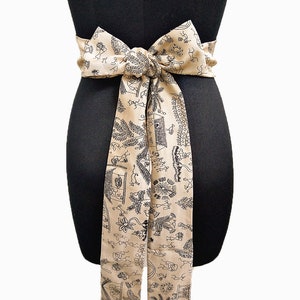 May include: A beige fabric sash tied in a bow, featuring a repeating black line art pattern of figures, plants, and structures. The sash is wrapped around a black mannequin, showcasing its design and drape.