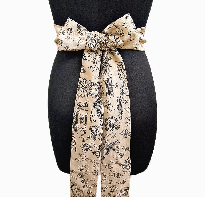 May include: A beige fabric sash tied in a bow, featuring a repeating black line art pattern of figures, plants, and structures. The sash is wrapped around a black mannequin, showcasing its design and drape.