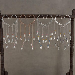 Könnte beinhalten: Vier Paare herzförmiger Ohrklemmen in Silber- und Roségoldtönen. Jede Klemme verfügt über mehrere hängende Ketten, die in kleinen metallischen Blumenanhängern und bunten Perlen enden und an einem geschwungenen Rahmen befestigt sind.